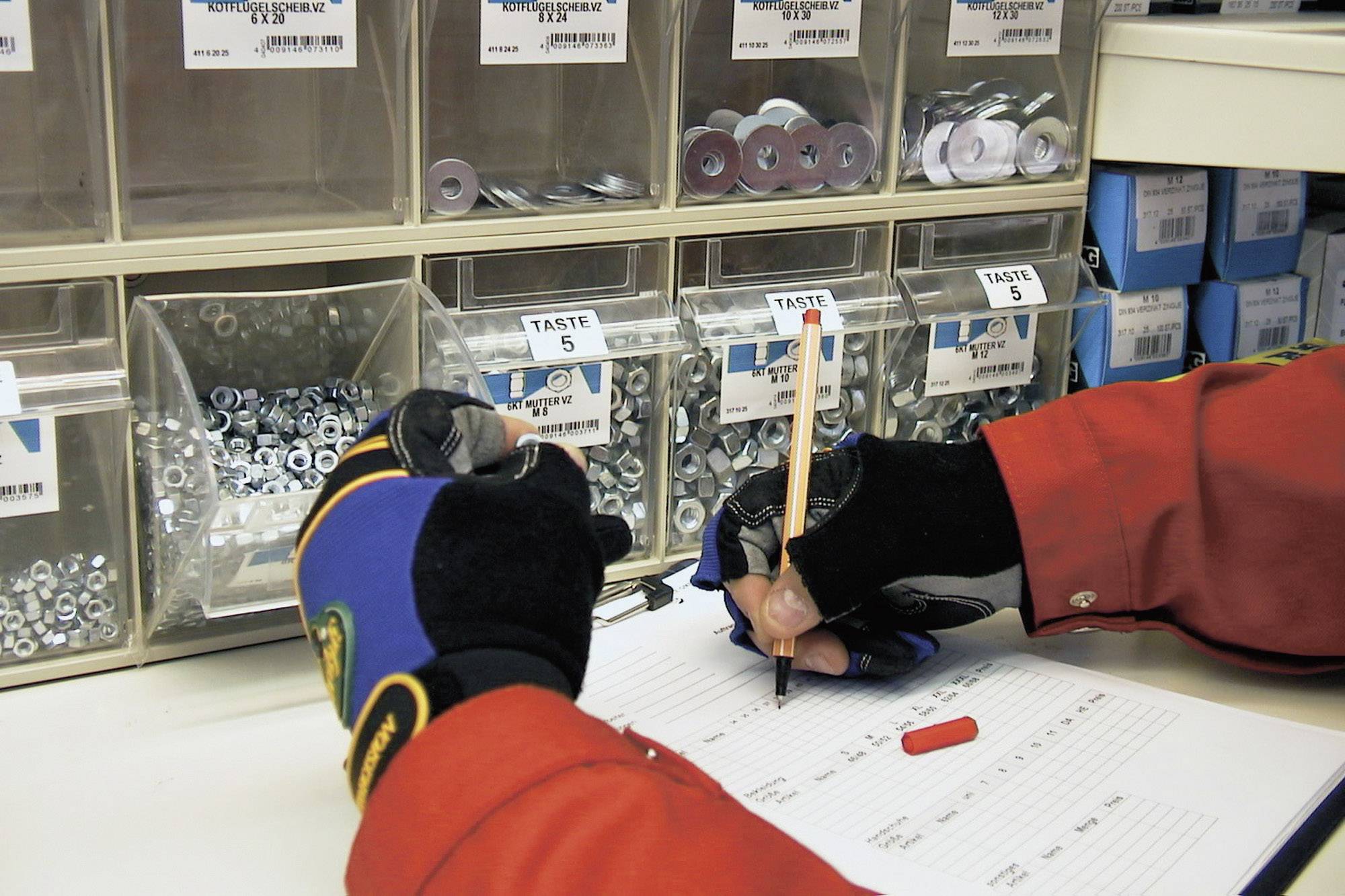 A person wearing gloves is writing with a pencil on a form in front of a shelf stacked with screws and nuts.