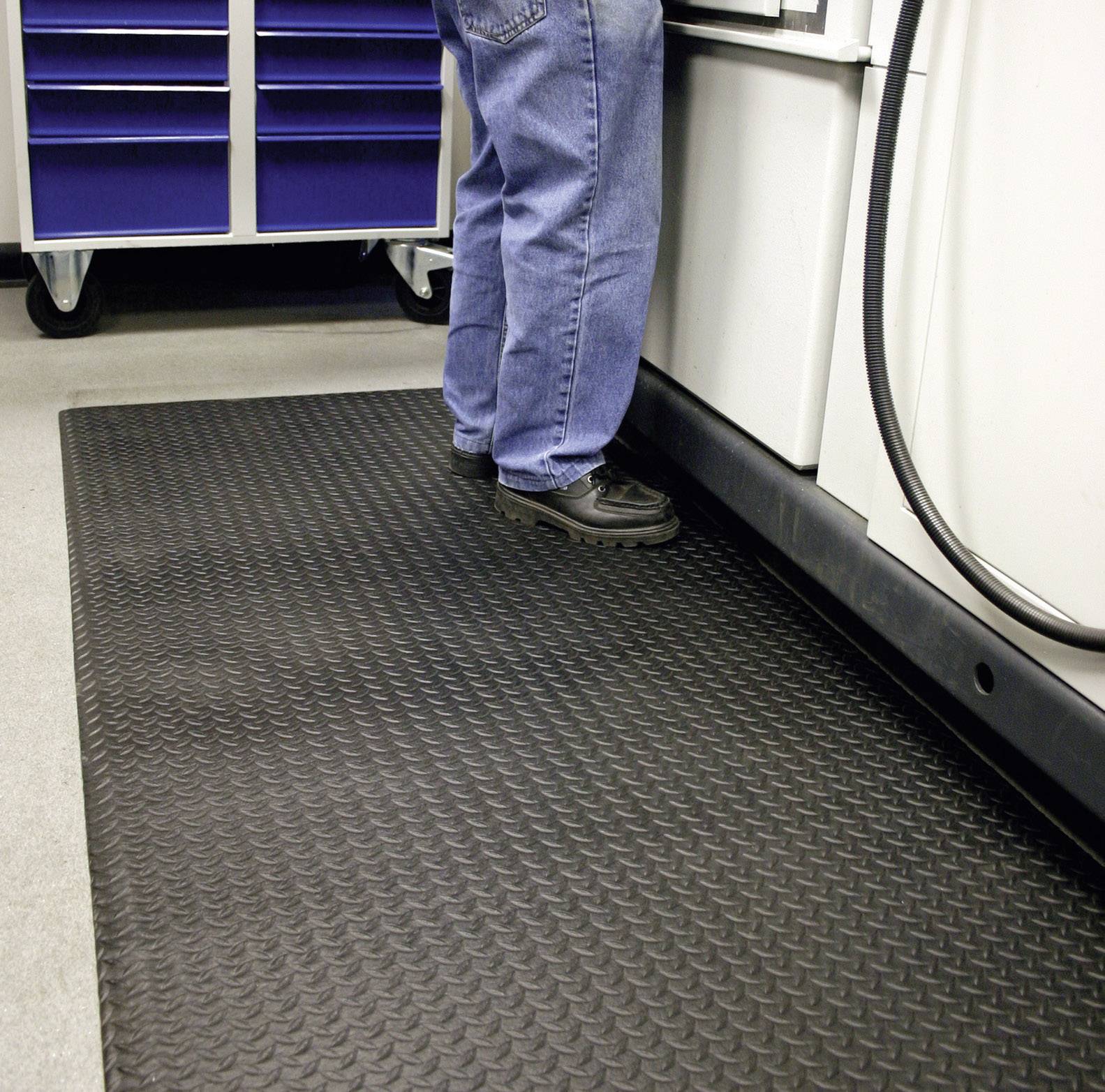 A person is standing on a black, textured floor mat in front of a cabinet with blue drawers in a workshop environment.