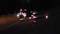 Two people in safety clothing are standing in front of a car at night. They are surrounded by traffic safety cones, possibly during an inspection.