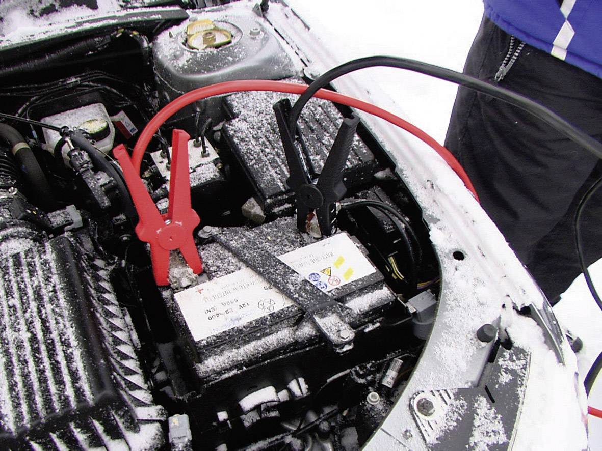 Car battery being jump-started with jump leads, red and black clamps connected. Wintry surroundings, snow visible.