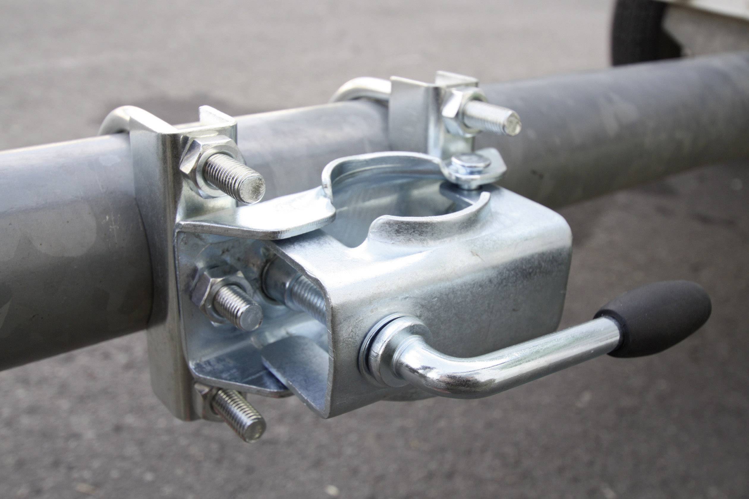 A metal clamp secures a pipe to a vehicle trailer, visibly fastened with bolts and a lever mechanism, on a grey asphalt surface.