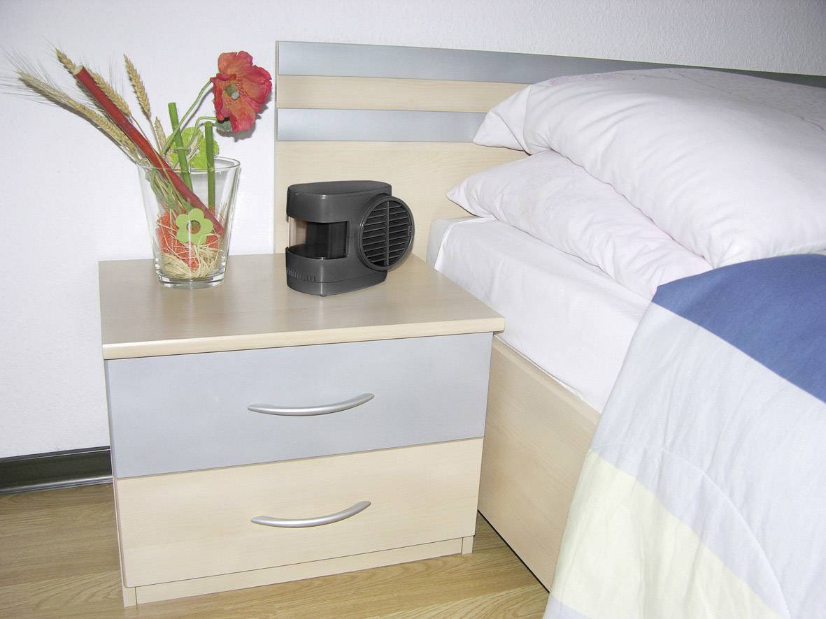 Bedside table with a glass vase full of flowers and a small black device beside it, next to a bed with a white-covered duvet.