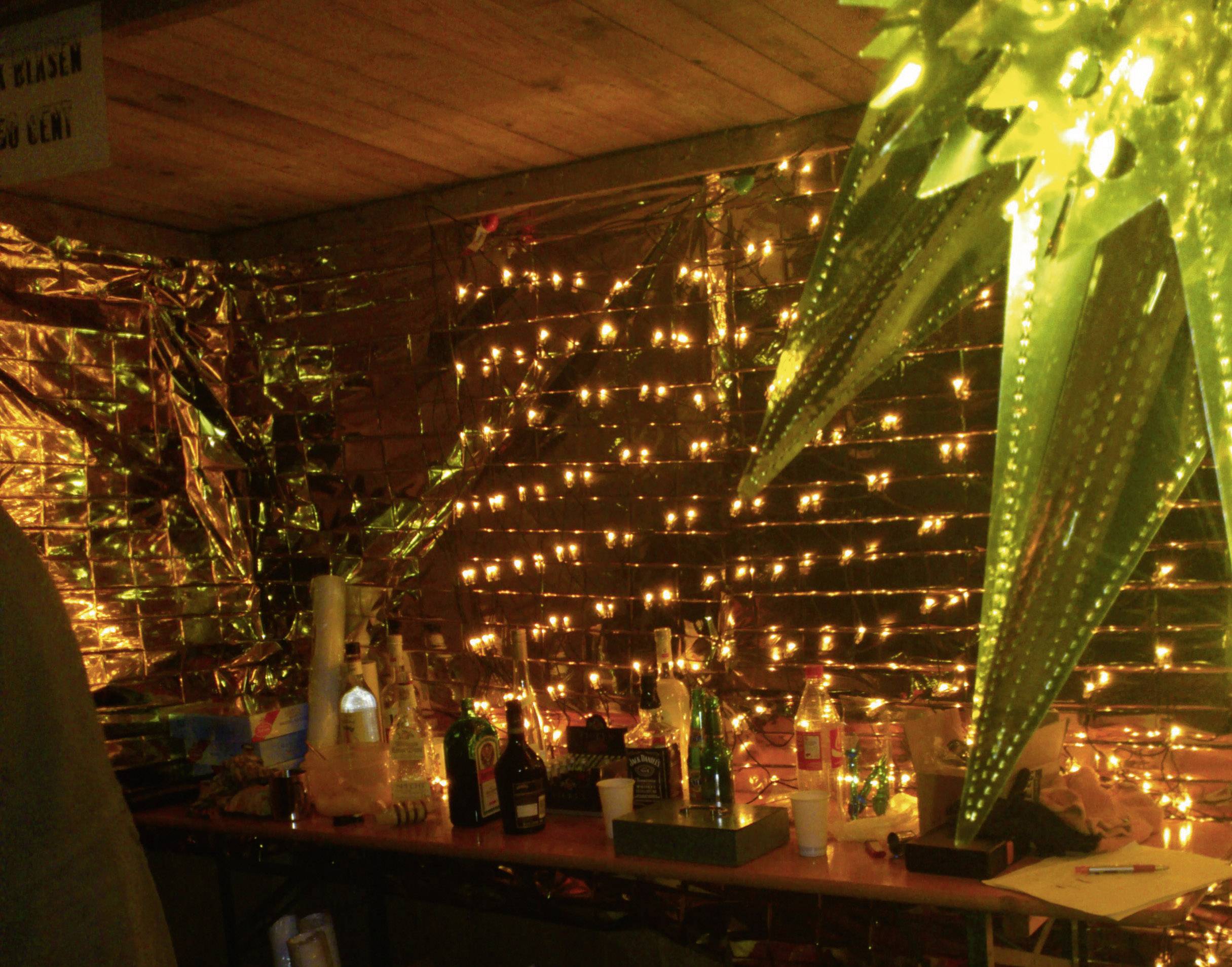 A festive bar with various bottles and glasses on a table. In the background, many small lights are glowing.