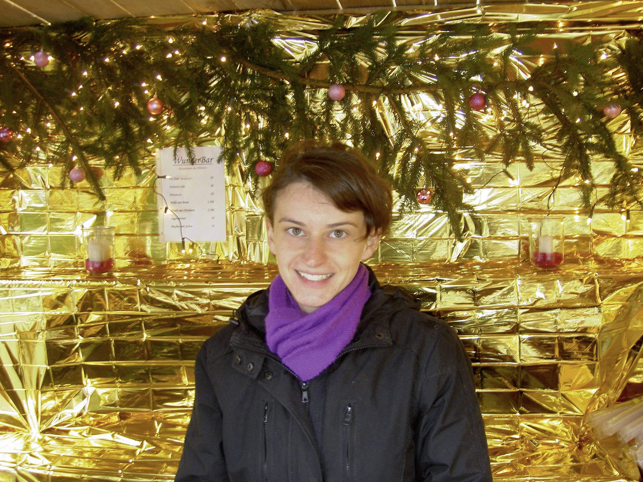 A person is smiling in front of a wall decorated with gold foil and pine branches. Lights and pink baubles are part of the decoration.