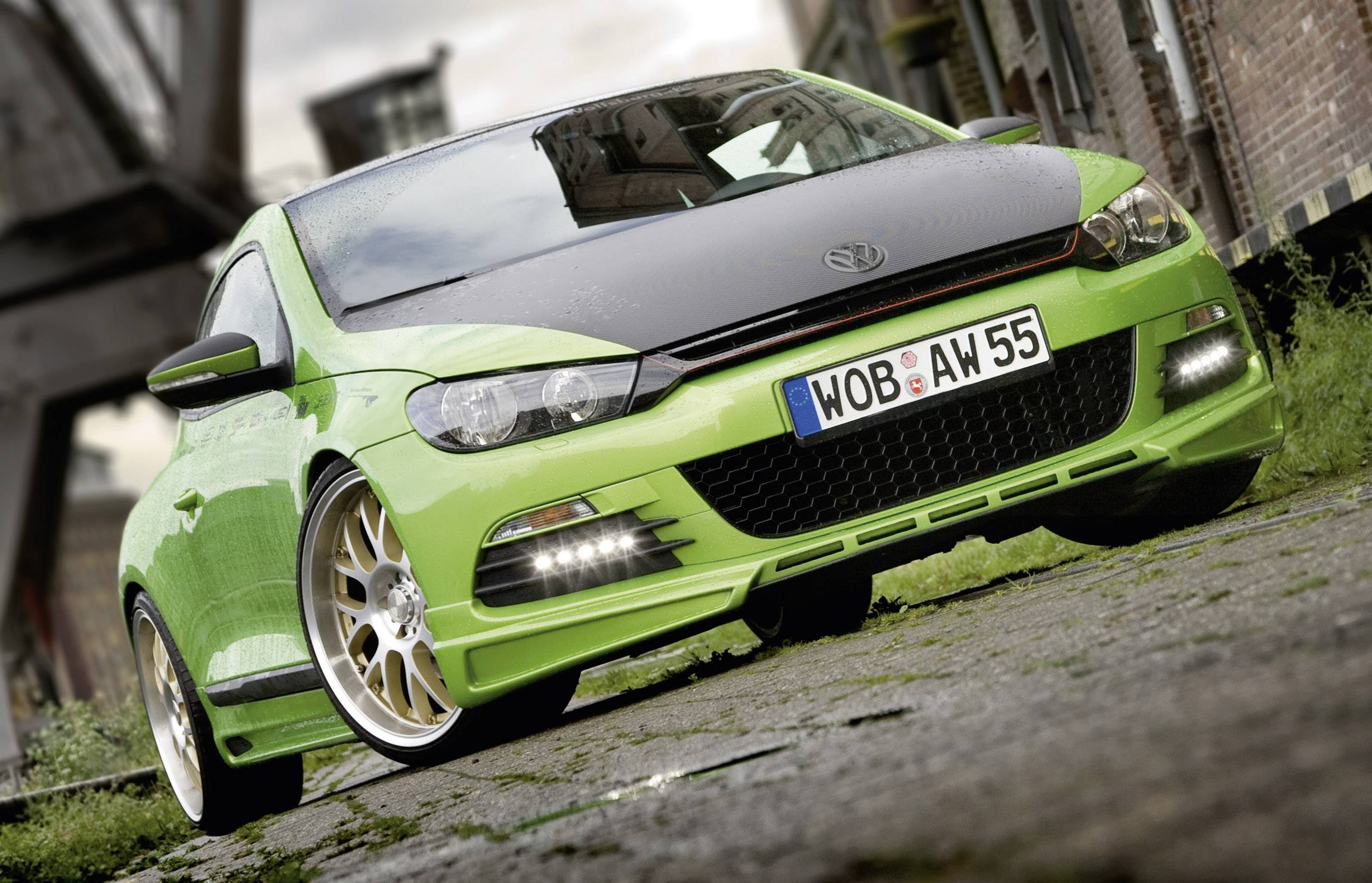 A light green sports car is parked on a cobbled path between brick buildings. It has a black bonnet and gold alloy wheels.