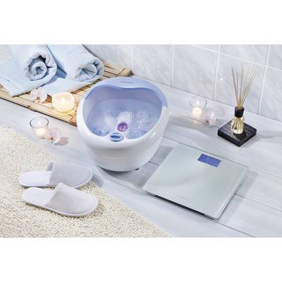 Foot bath with blue trim, filled with water and massage function. Surrounded by towels, candles, and a body scales on light wooden flooring.