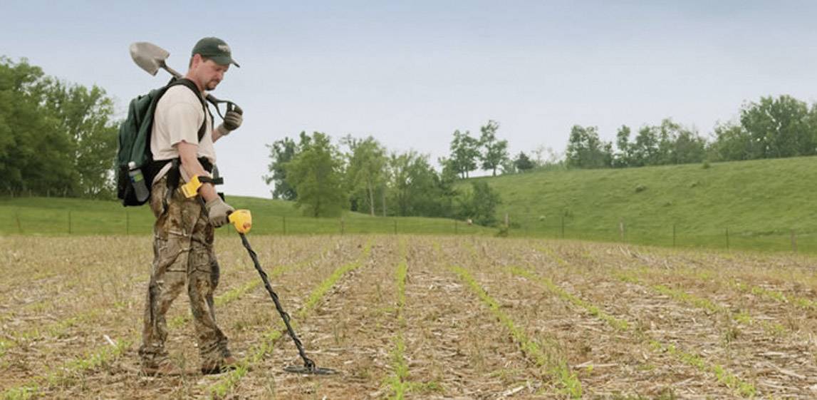 A person with a spade and metal detector is searching for hidden objects on a large, open field. Trees and a hill can be seen in the background.