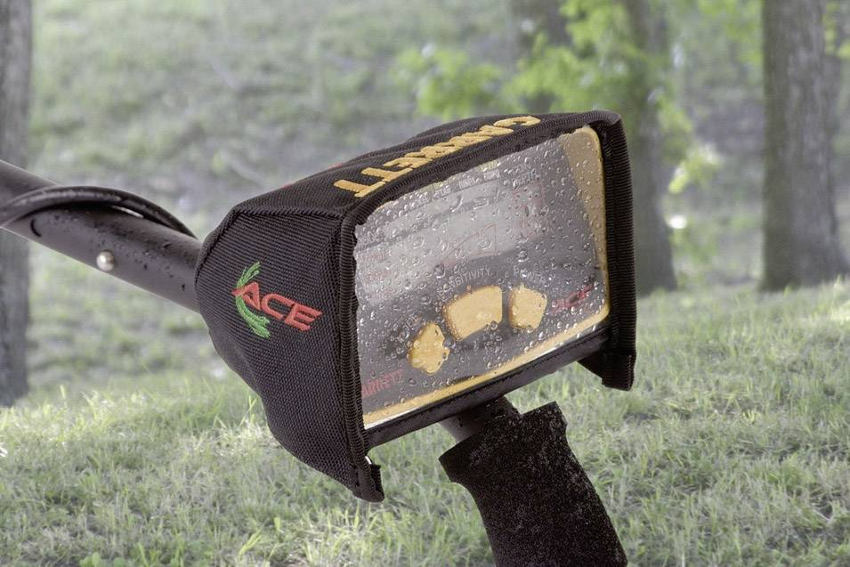 A metal detector with a rain-soaked cover displays a screen. The background is a green, wooded landscape.