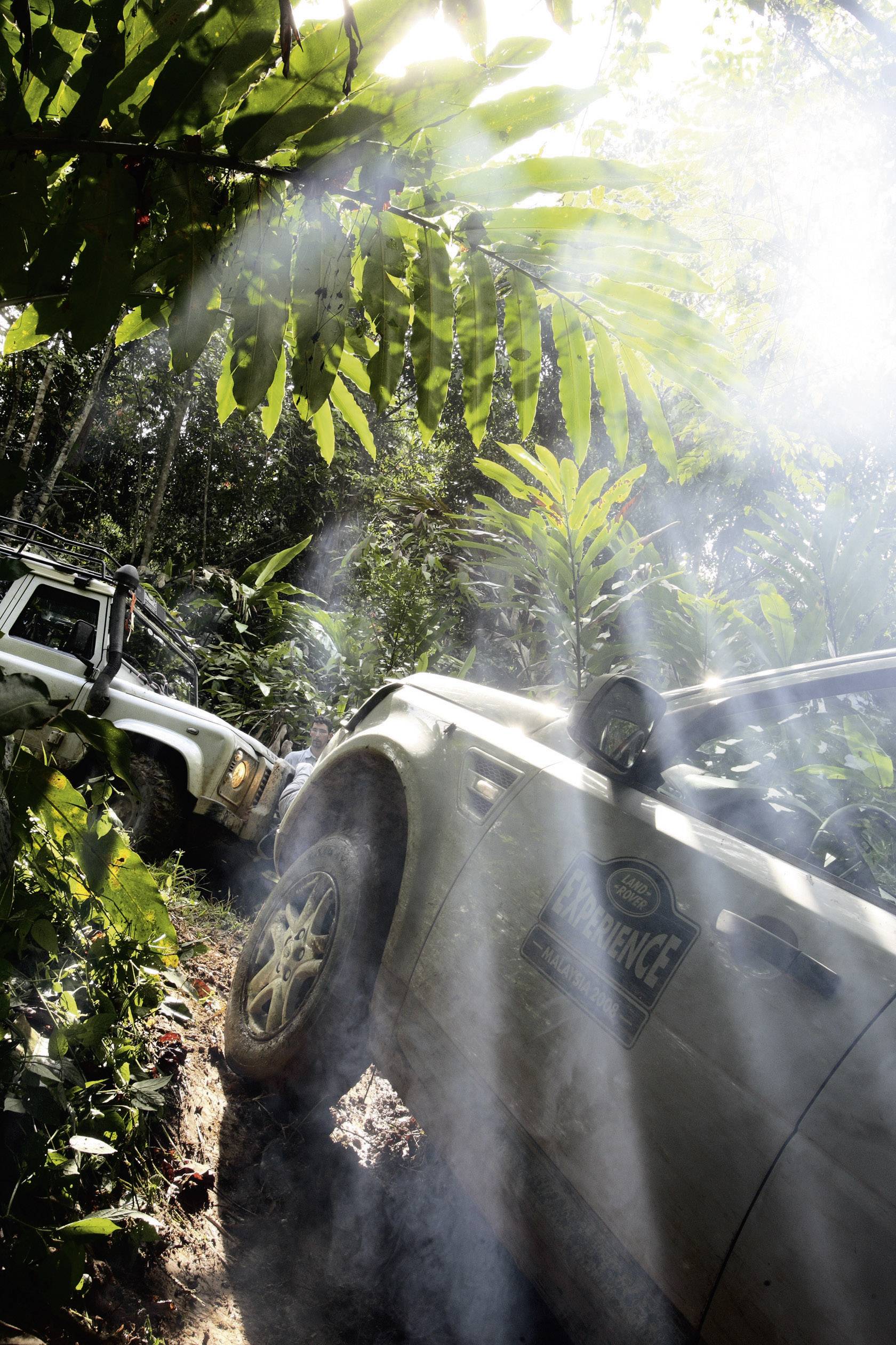 Two off-road vehicles drive along a muddy, sunlight-drenched jungle track, surrounded by dense vegetation.
