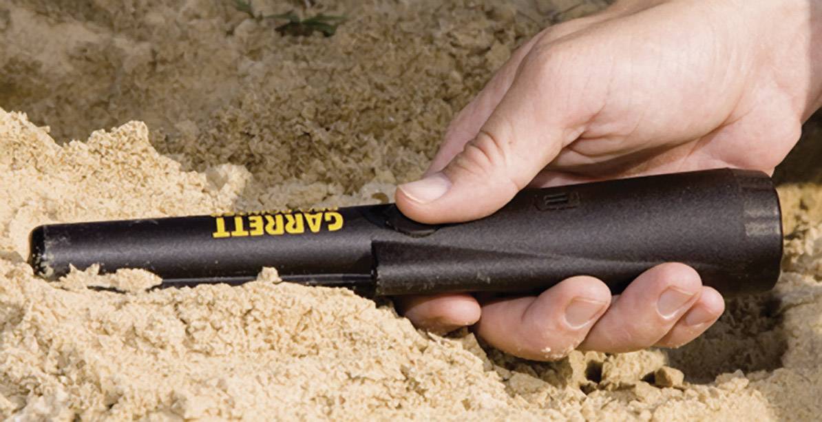 A hand is holding a black metal detector with the label 'Garrett', which is positioned in sandy soil.