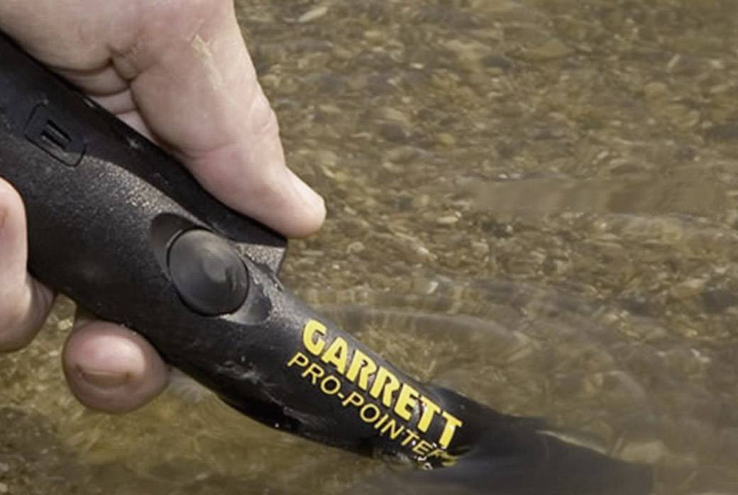 A hand is holding a black metal detector called the 'Garrett Pro-Pointer', partially submerged in water, over a gravelly surface.