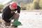A person wearing a red helmet is searching for gold nuggets along the bank of a shallow river. Wooded hills can be seen in the background.