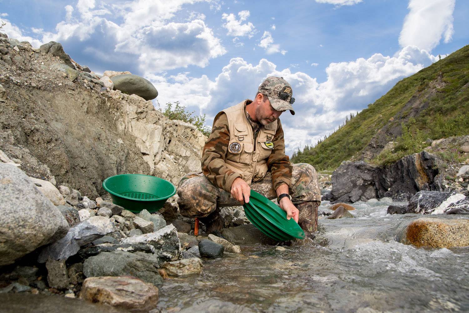 A man in camouflage clothing sits beside a mountain stream, washing gold with a green gold pan. Cloudy sky in the background.