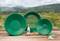 Three green gold panning dishes and accessories on a wooden table against a forested landscape, used for gold prospecting in rivers.