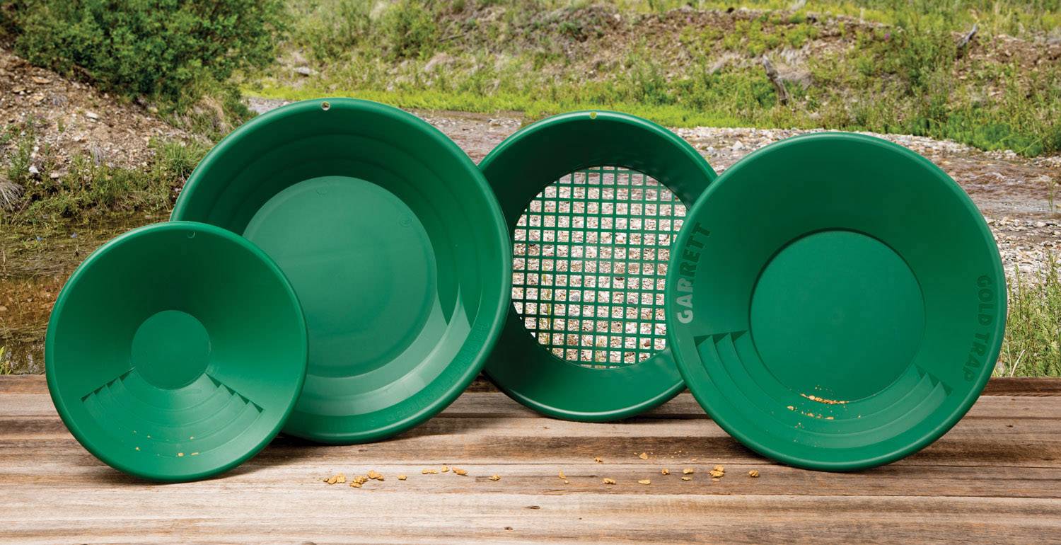 Four green gold-panning trays on a wooden table outdoors. Some show small gold flakes, with a forested background.