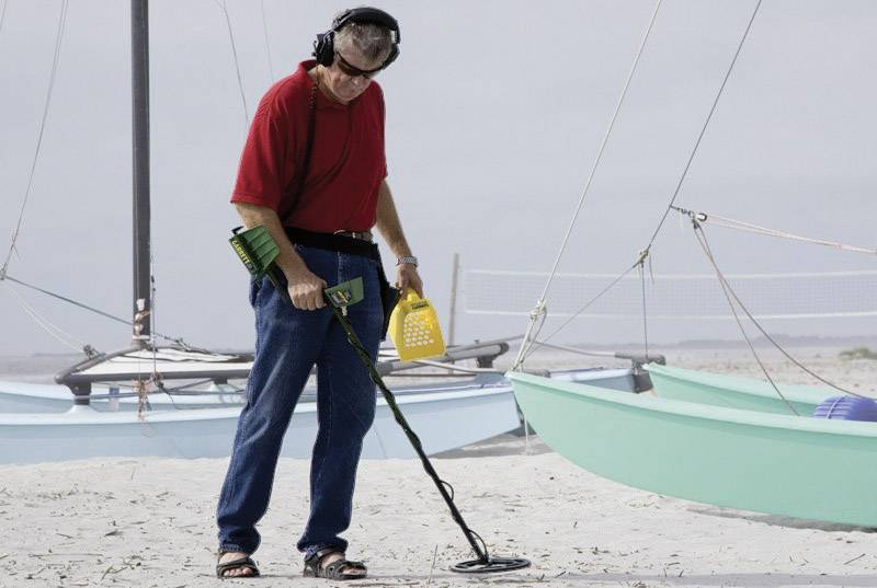 A person is searching with a metal detector on the beach, surrounded by sailing boats.