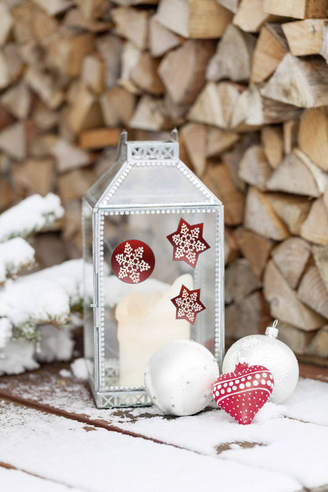 A lantern with a candle, decorated with red and white Christmas stars, stands on a wooden table in the snow in front of stacked firewood.