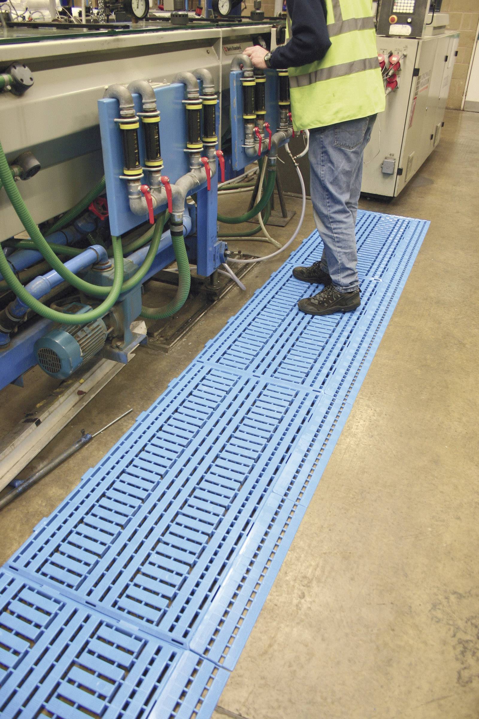 A person is working on a machine in a factory environment. Blue floor mats are visible, covering the work area.
