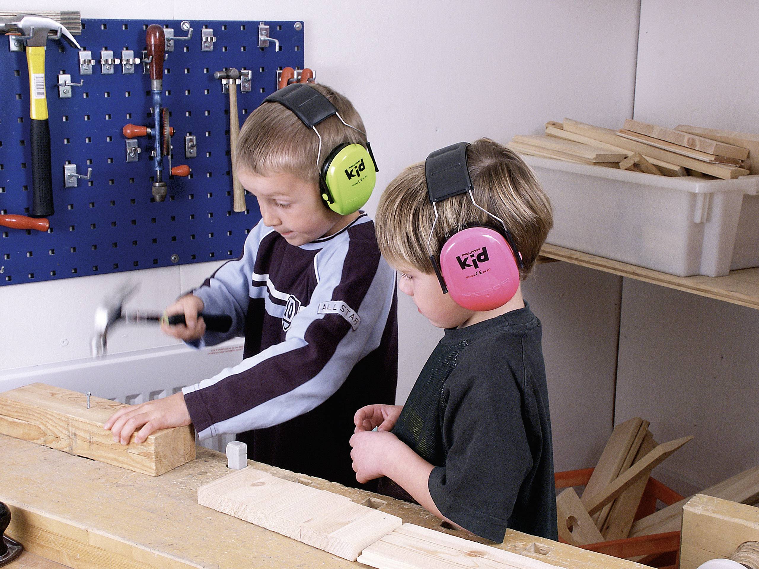 Two children wearing ear defenders are working in a workshop. One is hammering a nail into a piece of wood, while the other looks on.