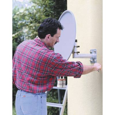 A man in a checked shirt is installing a satellite dish on a yellow wall. Plants can be seen in the background.