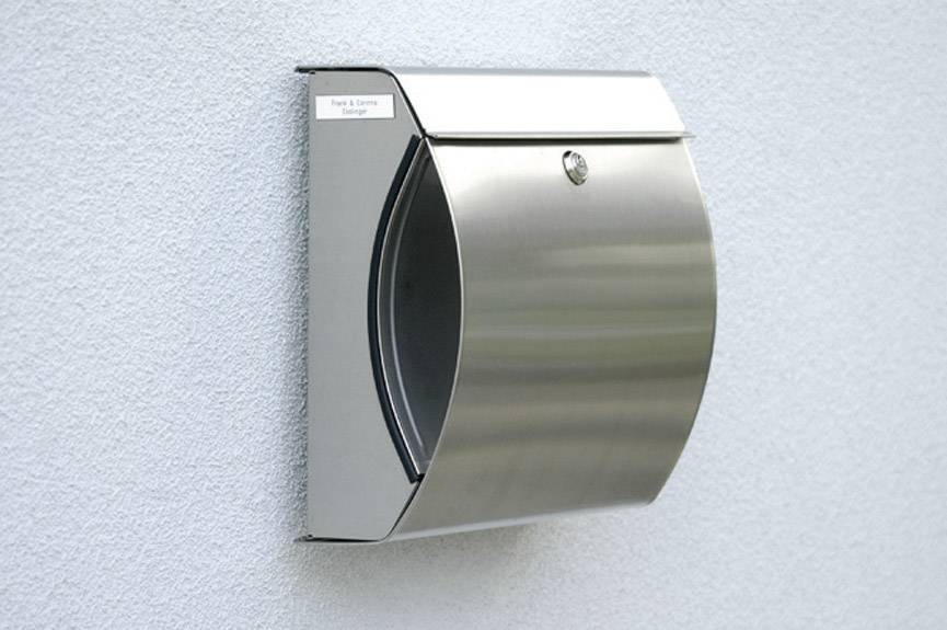 Modern, brushed stainless steel letterbox mounted on a wall. Minimalist, contemporary design with a front flap and small name strip in the top left corner.