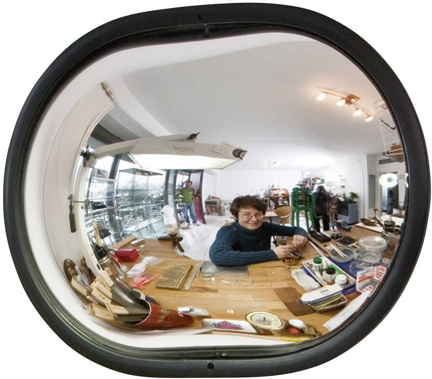 A woman sits in a room with model-making accessories. The environment is reflected in an oval, convex mirror.