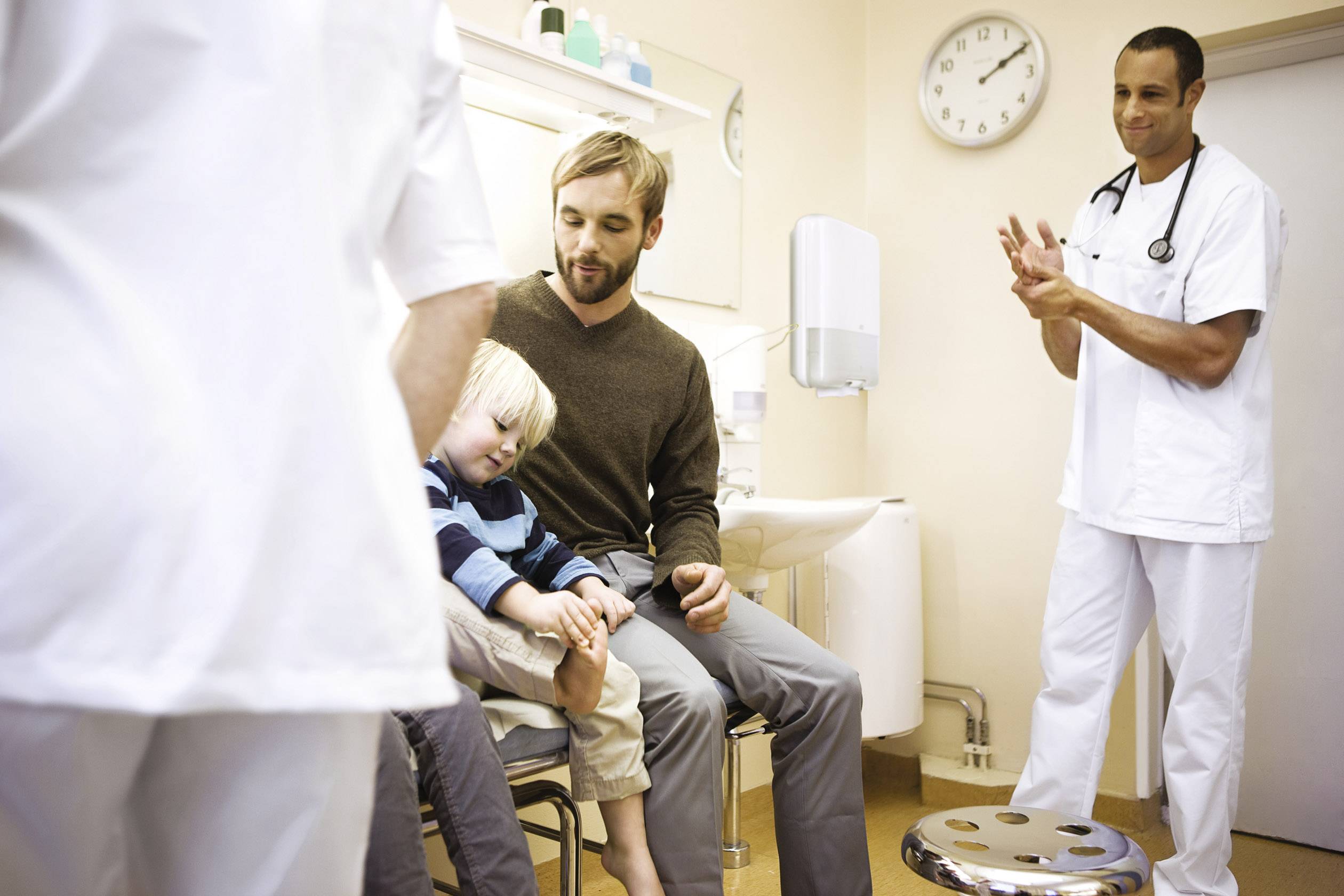 A Doctor's Appointment: A man sits with a small child in an examination room, while a doctor stands nearby and observes.