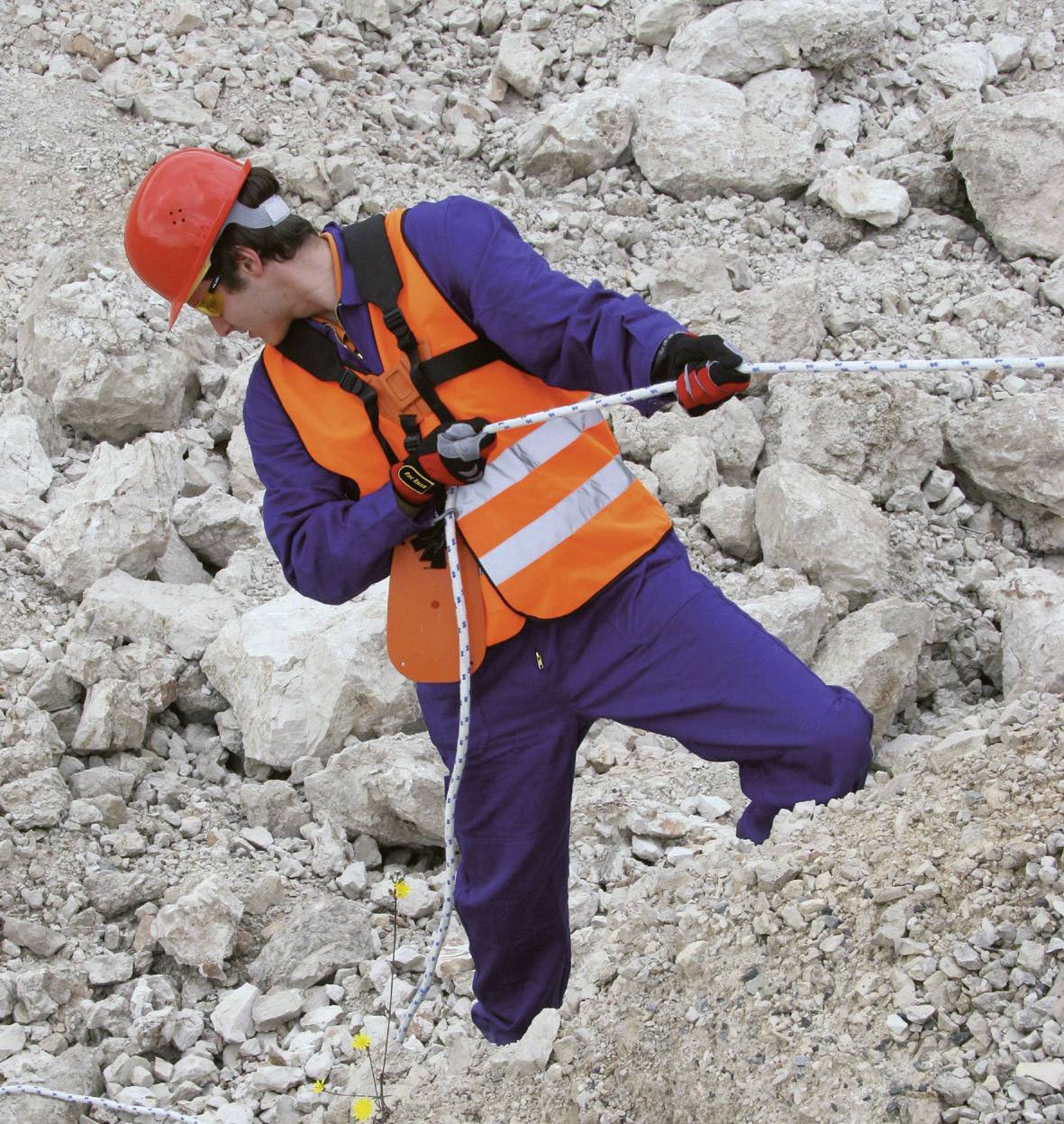 A person in protective clothing and a helmet is holding a rope while climbing down a rocky slope.