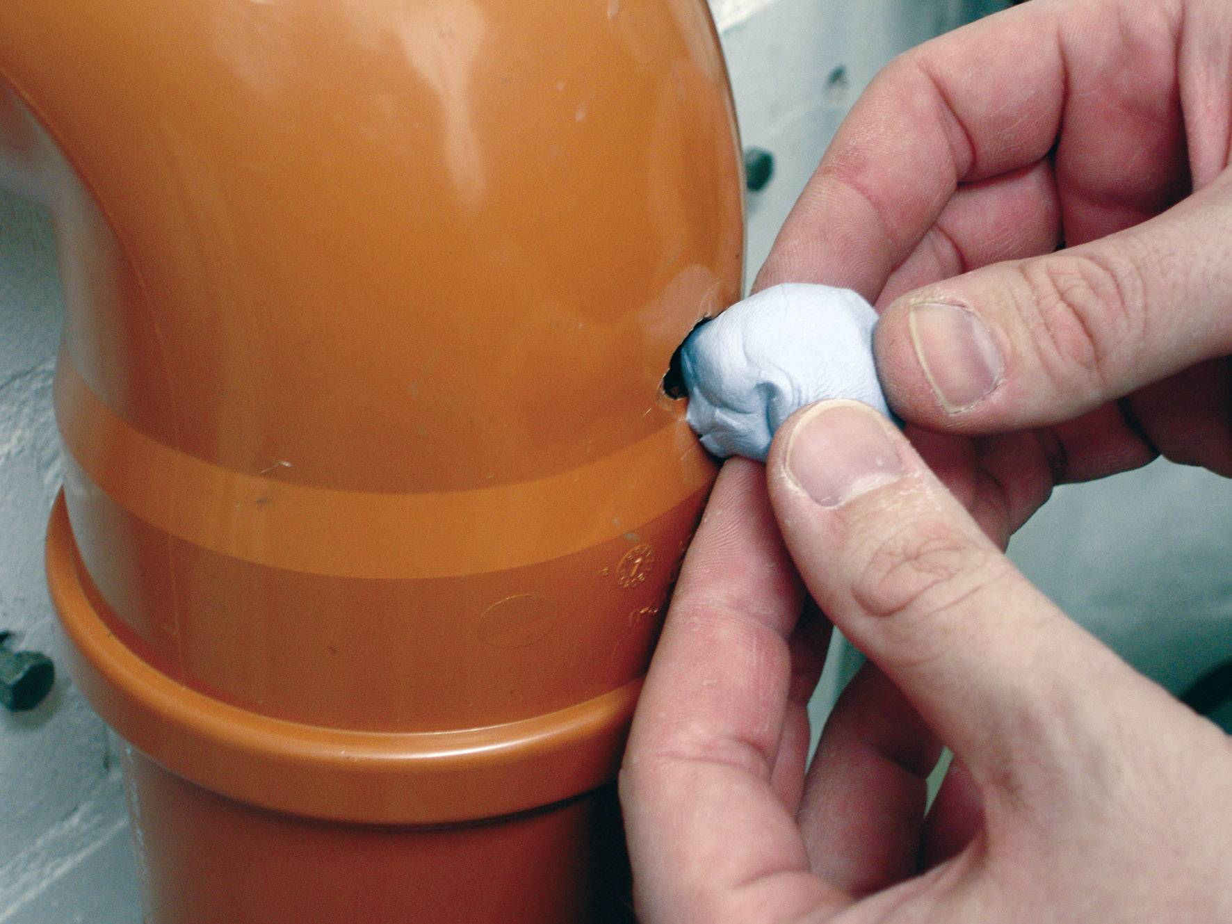 A person is repairing a small hole in an orange pipe with blue putty to prevent a leak.