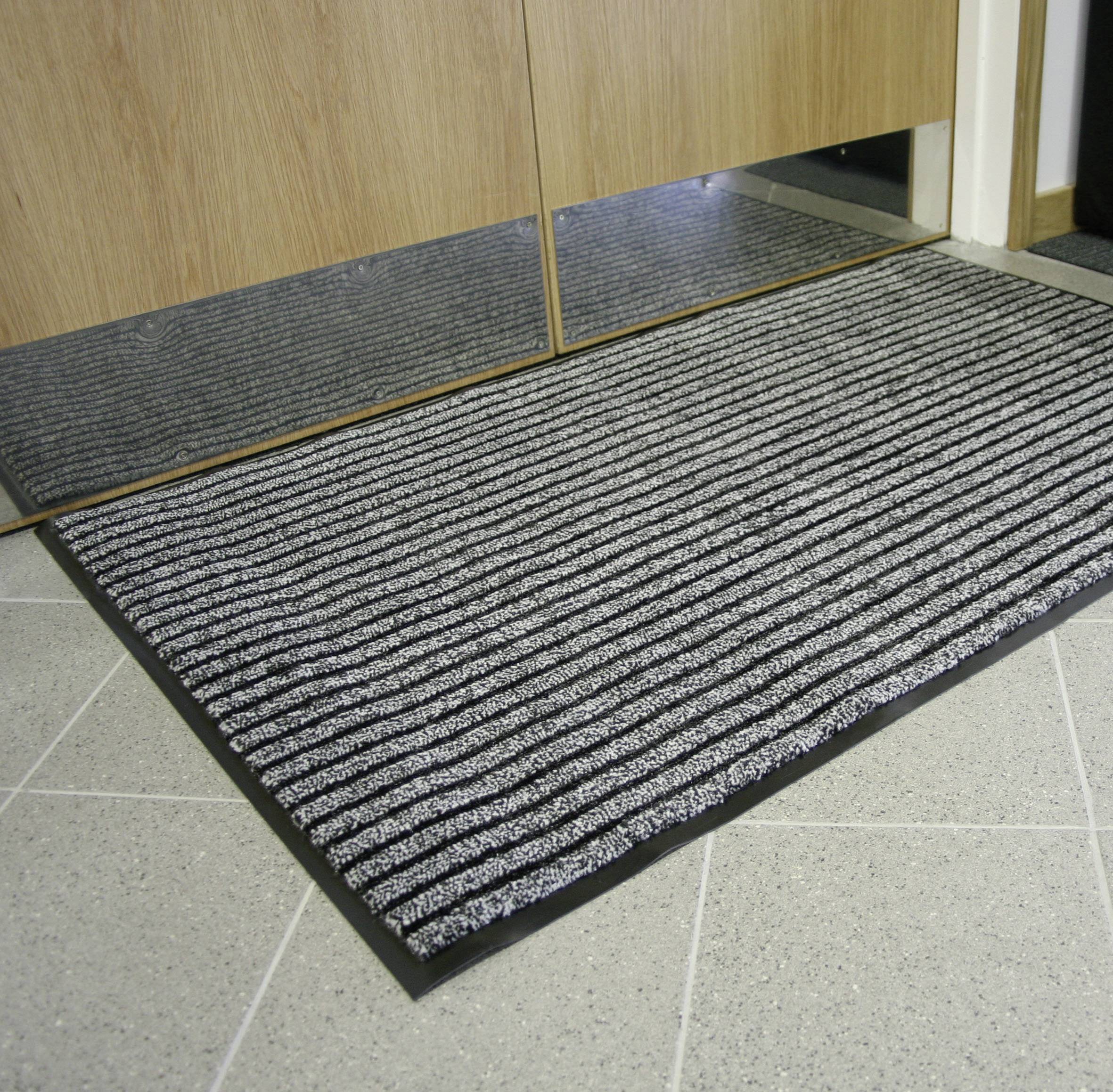 A grey doormat with black stripes lies in front of a wooden door on a tiled floor.