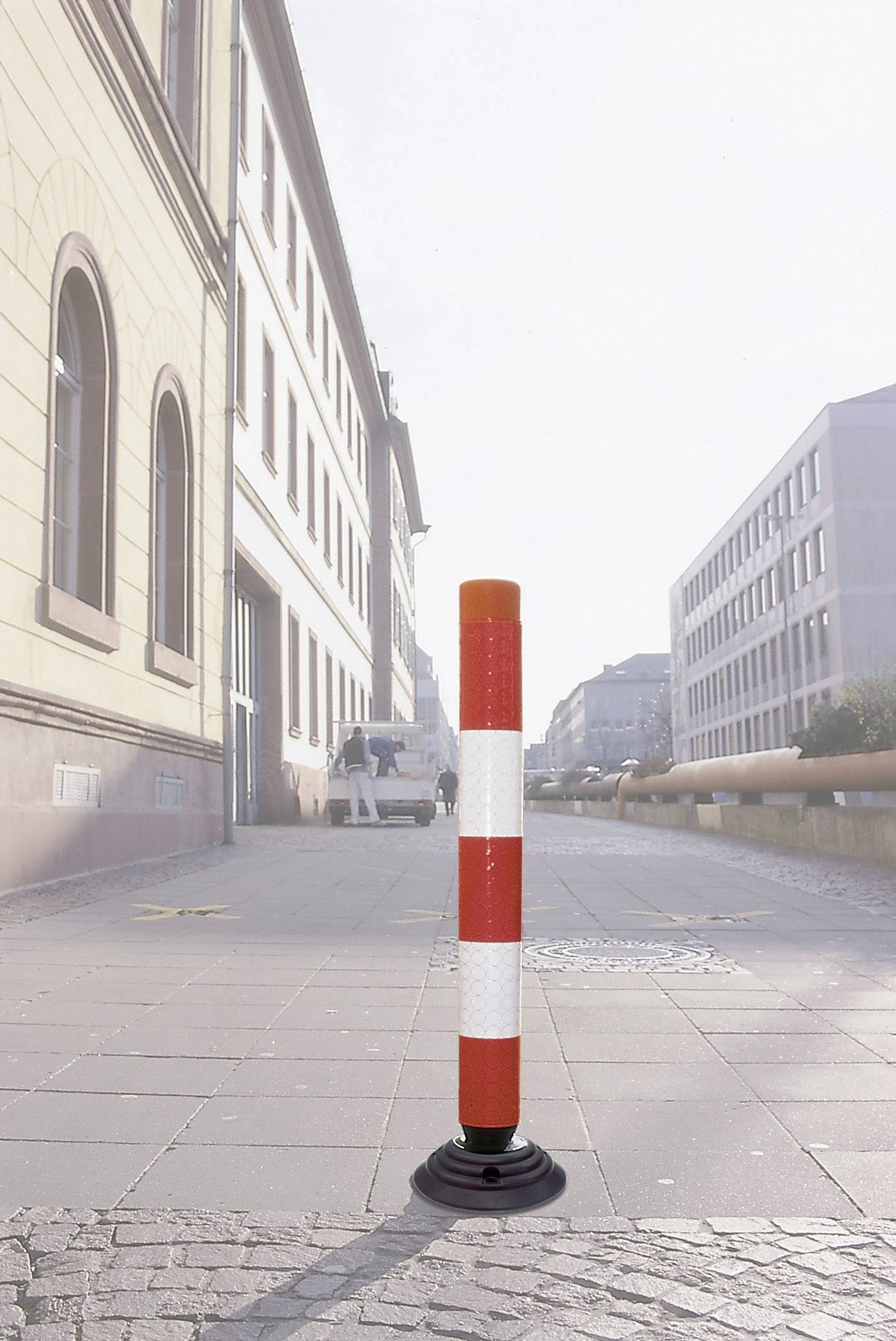 A red and white striped barrier post stands on a paved pavement in an urban setting on a sunny day.