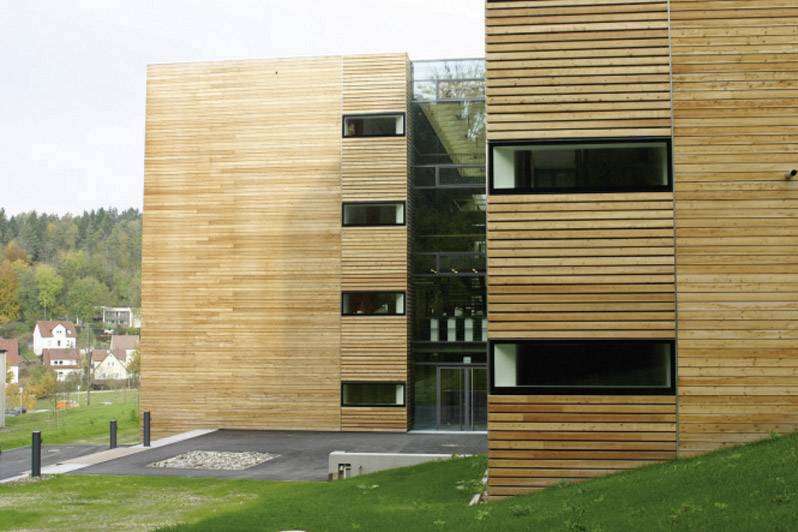 Modern building with wooden cladding, large glass windows, and a flat roof. Green lawn in the foreground, forest in the background.