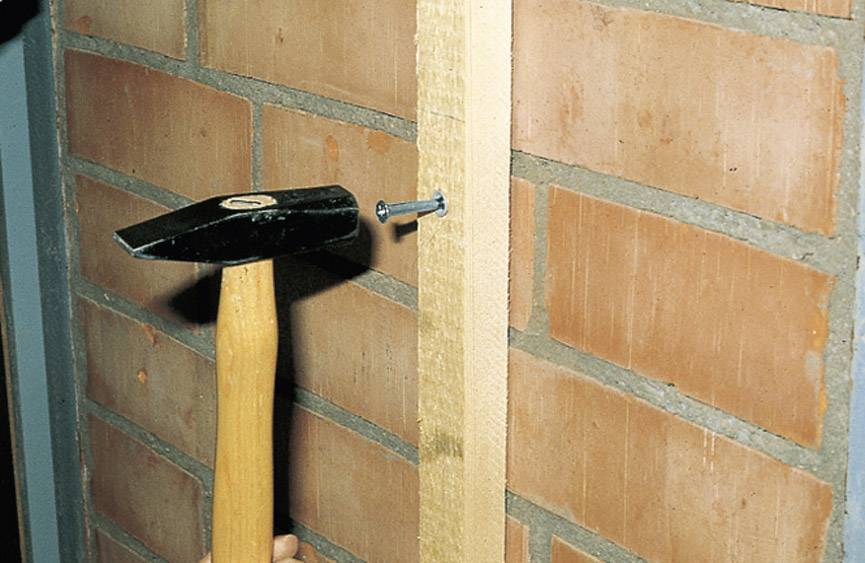 A hammer strikes a nail into a wooden board that is secured to a brick wall.