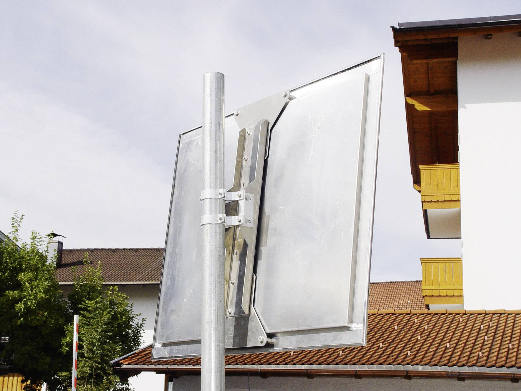 A metal frame for solar panels, mounted at an angle on a pole, with a house in the background beneath a cloudy sky.