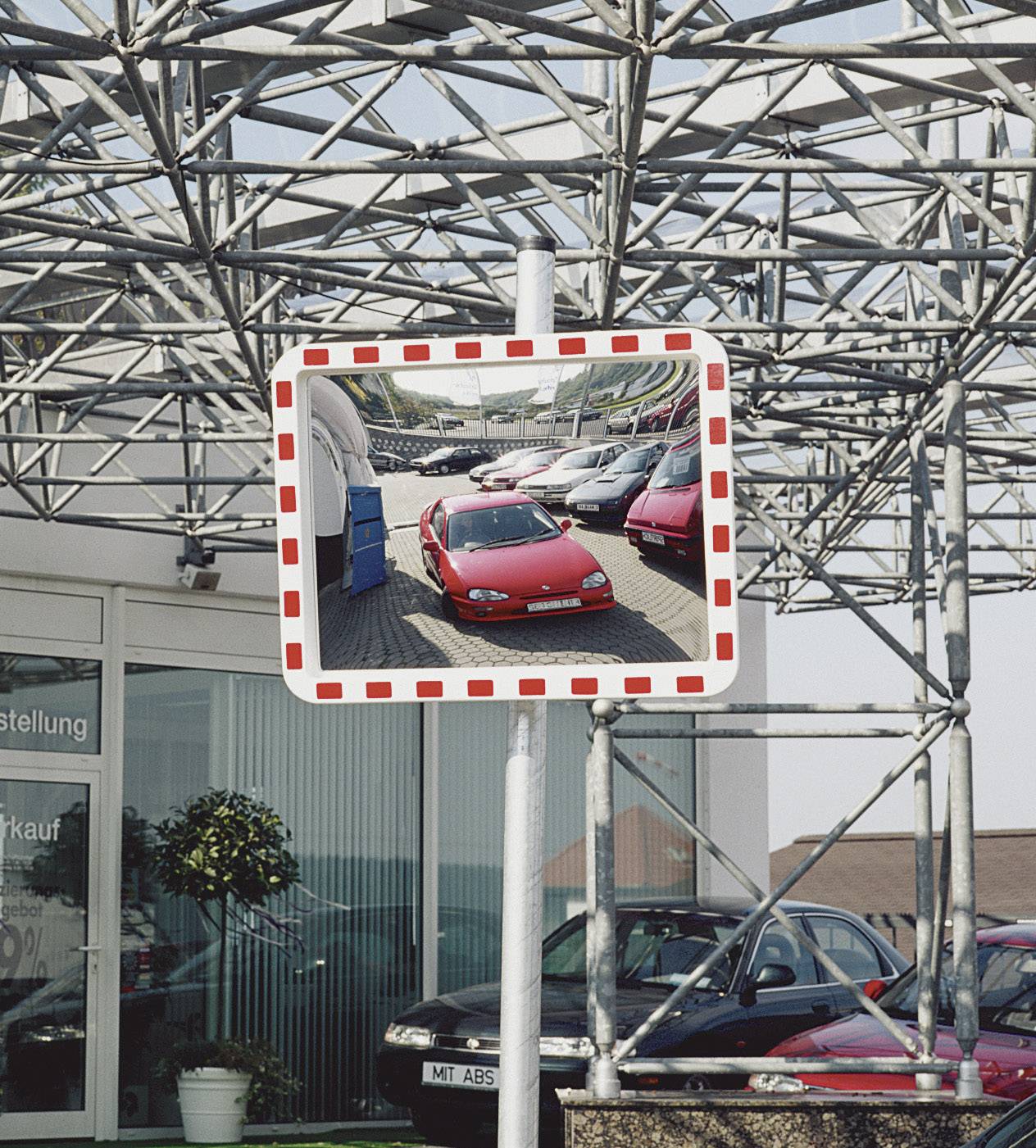 Traffic mirror beneath a scaffolding, showing parked red cars in a car park in front of a building.