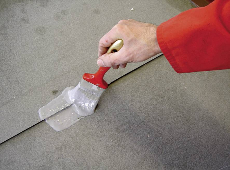 A hand holding a red tassel is applying a grey liquid to a wall joint. The person is wearing red clothing.