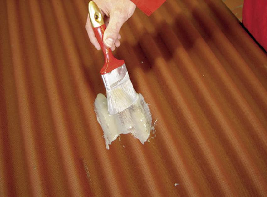 A hand applies white glue with a flat brush onto a wavy, brown surface.