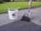 A person is spreading asphalt repair compound on a pavement with a shovel. Next to them is a white bucket with the label 'SAFE STEP'.