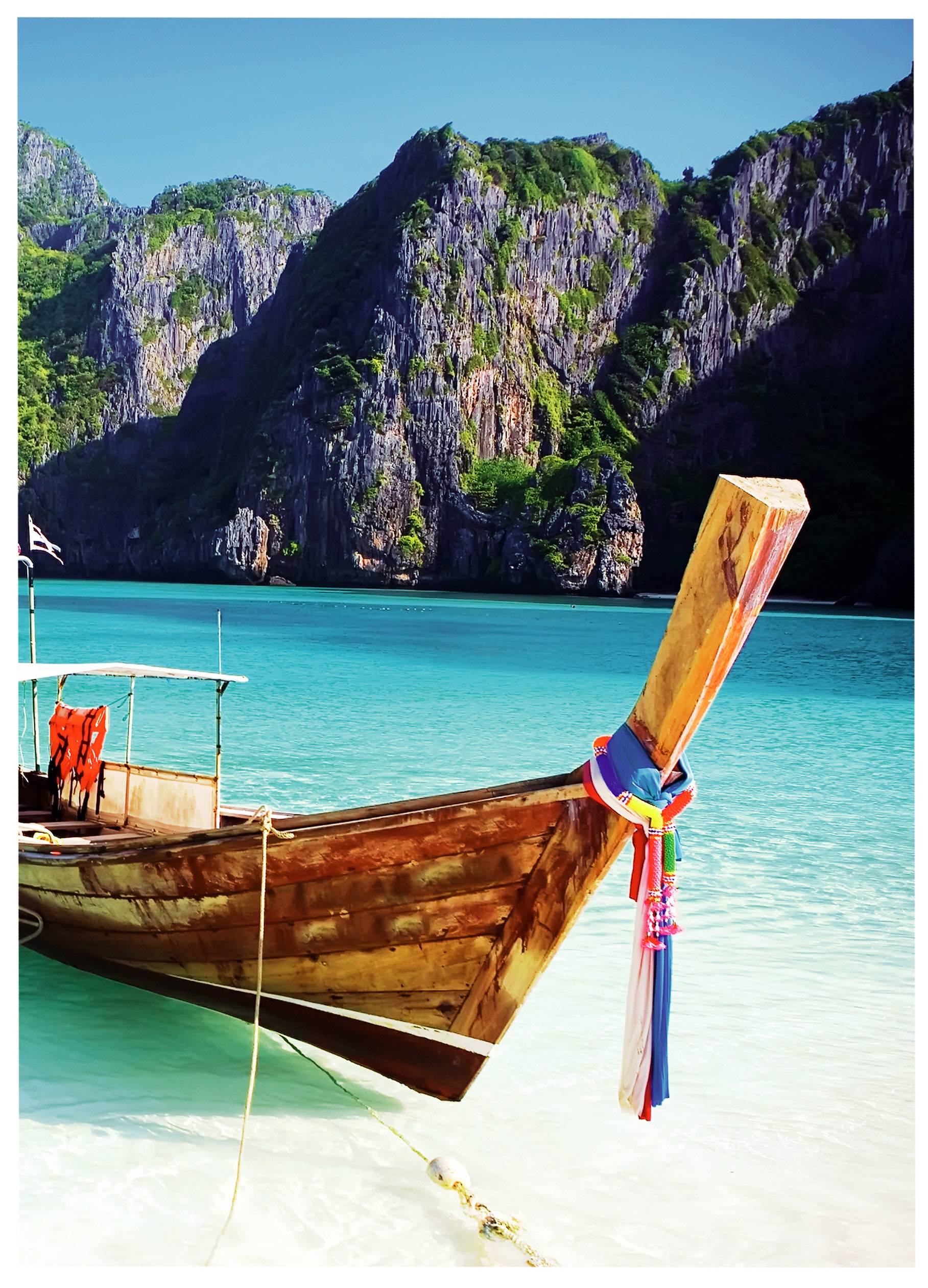 A wooden boat with colourful decoration anchors in turquoise water before steep, wooded cliffs under a clear sky.