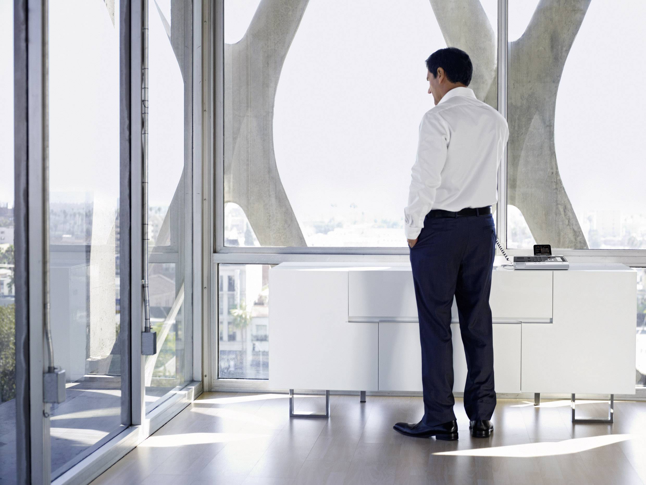 A man in business attire stands in a modern office in front of a large glass wall, gazing out at the cityscape beyond.