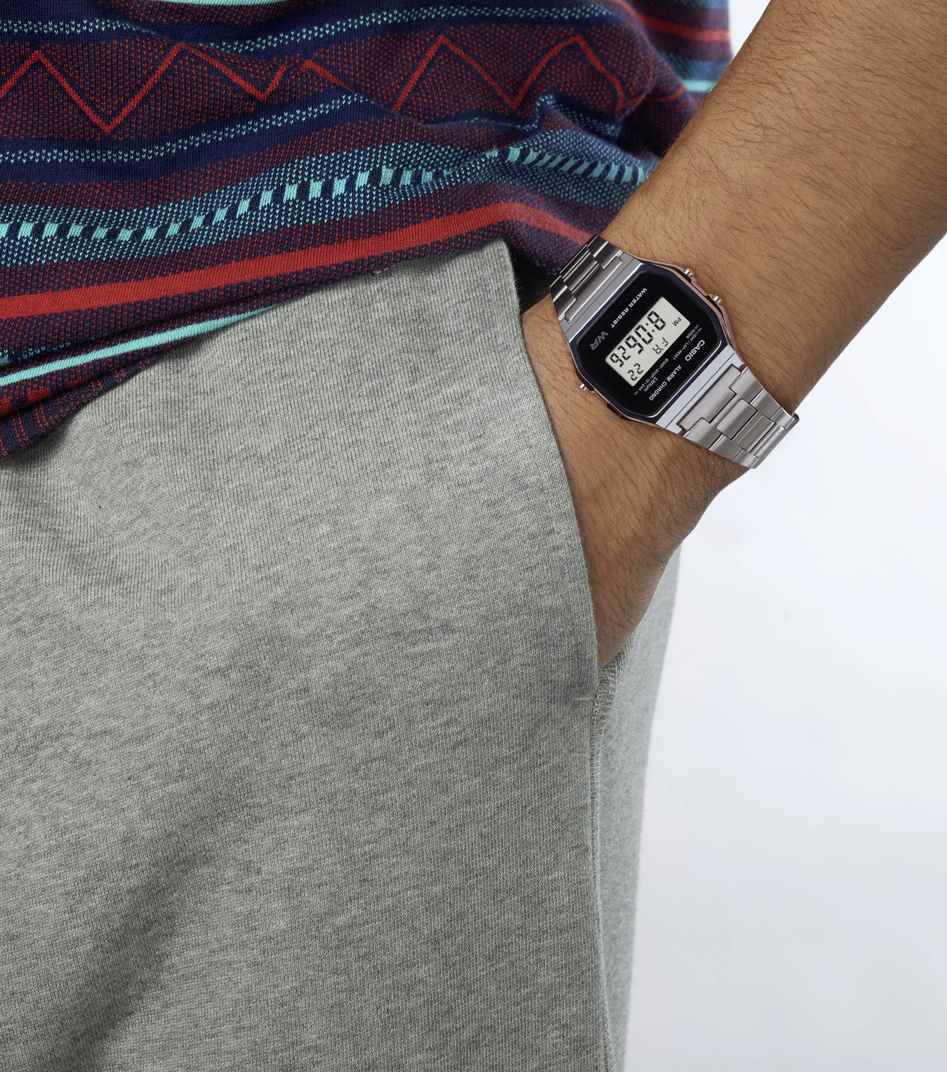 Personne portant une chemise à motifs, un pantalon gris et une montre-bracelet numérique dont l'heure est bien visible. Main dans la poche du pantalon.