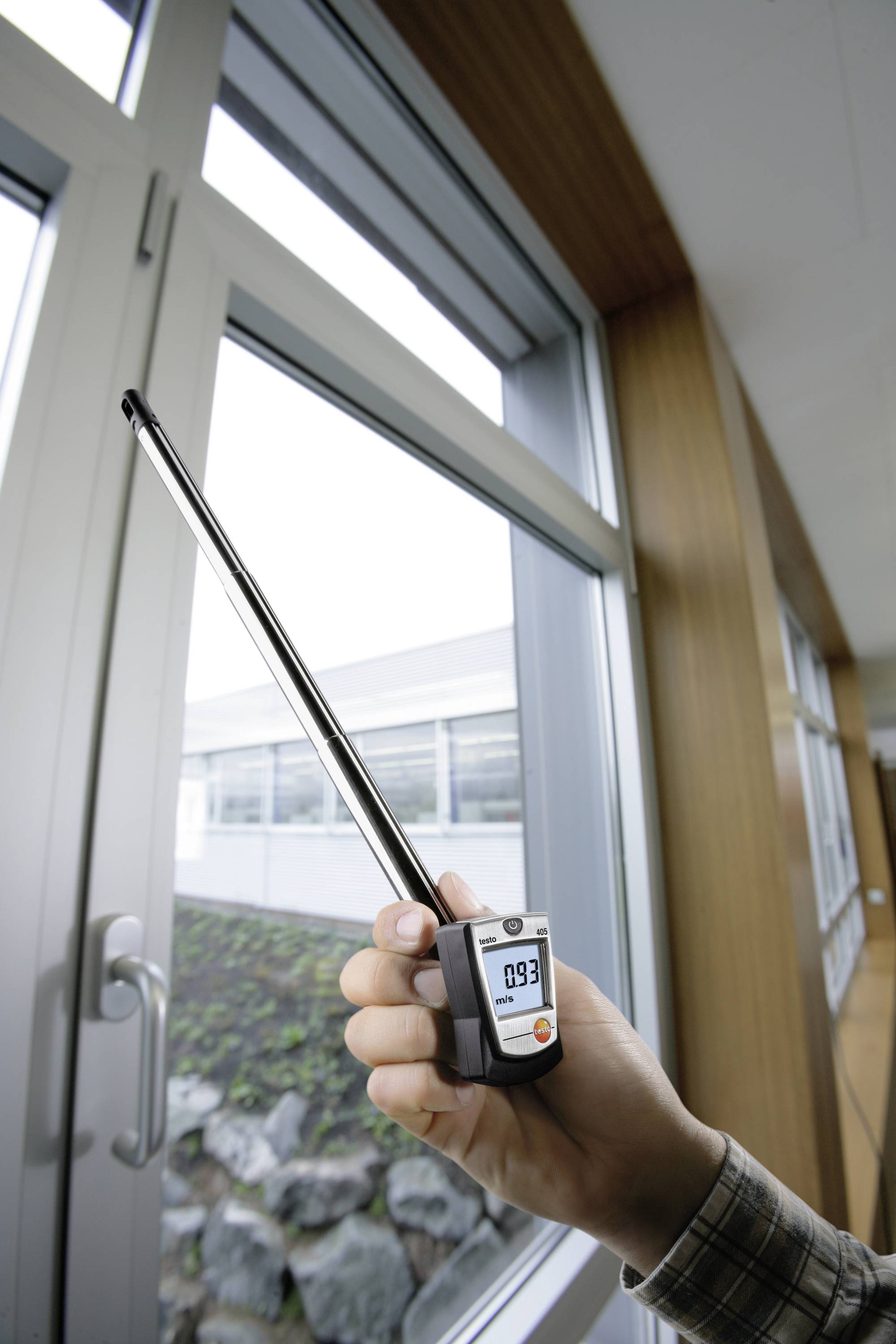 Une main tient un appareil de mesure électronique avec une longue sonde devant une fenêtre. L'affichage indique '0,9'.