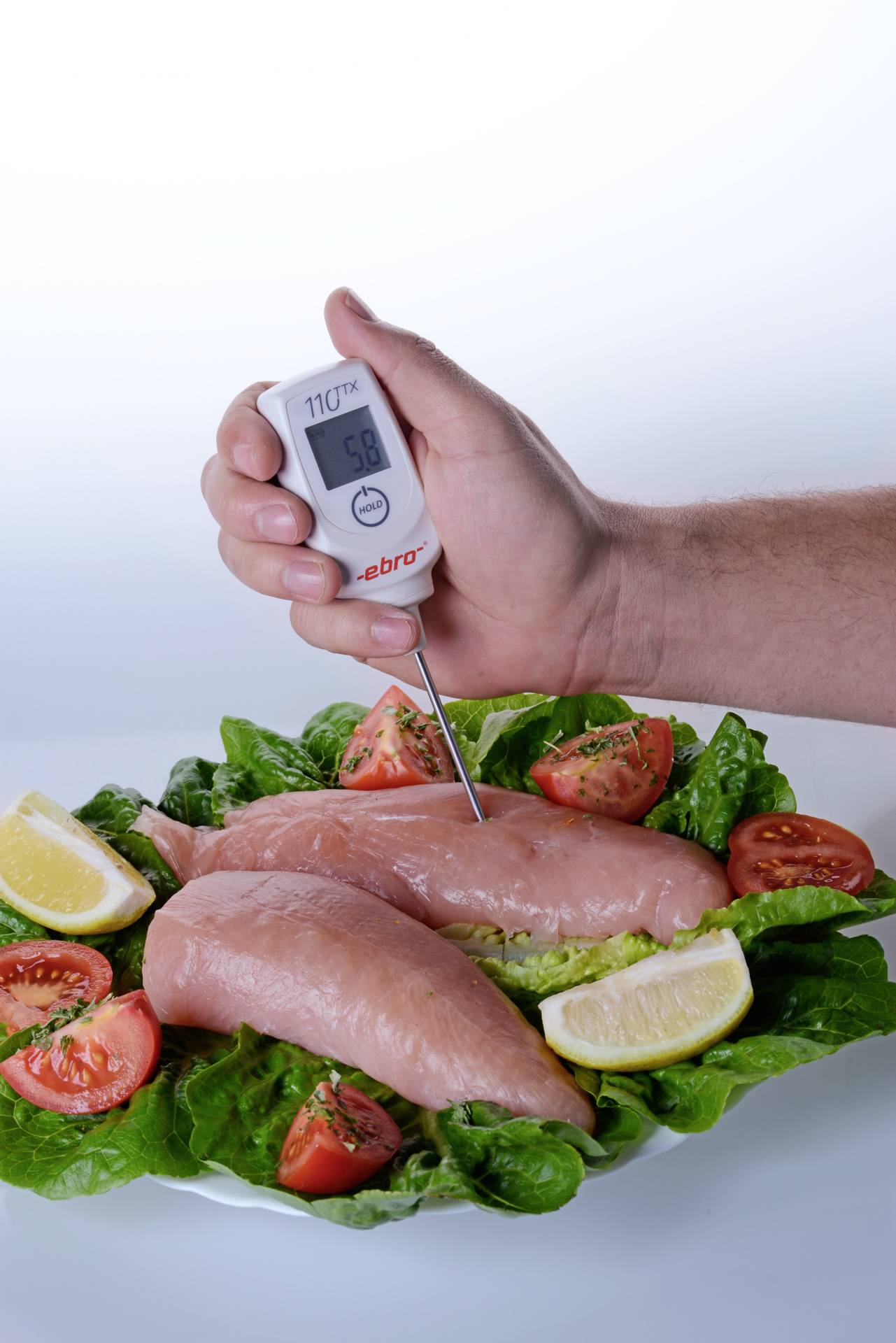 Un thermomètre de cuisine est inséré dans un poulet cru posé sur un lit de salade avec des tomates et des tranches de citron pour vérifier sa température.