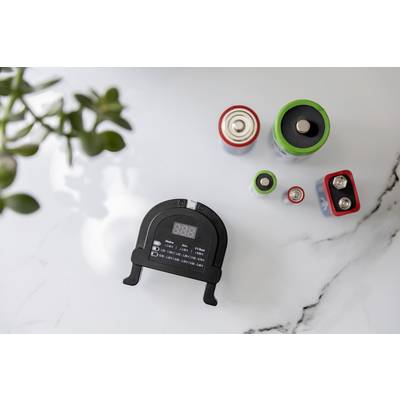 Un appareil de mesure de batterie noir est posé sur une table, entouré de différentes batteries. Des feuilles floues sont visibles à gauche.