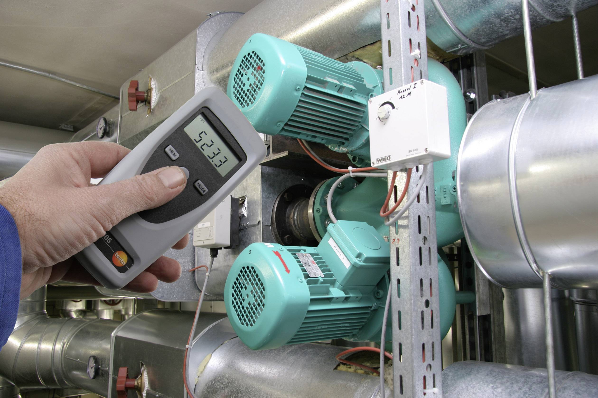 Une personne tient un appareil de mesure devant deux moteurs électriques verts dans un local technique, probablement pour vérifier leur bon fonctionnement.