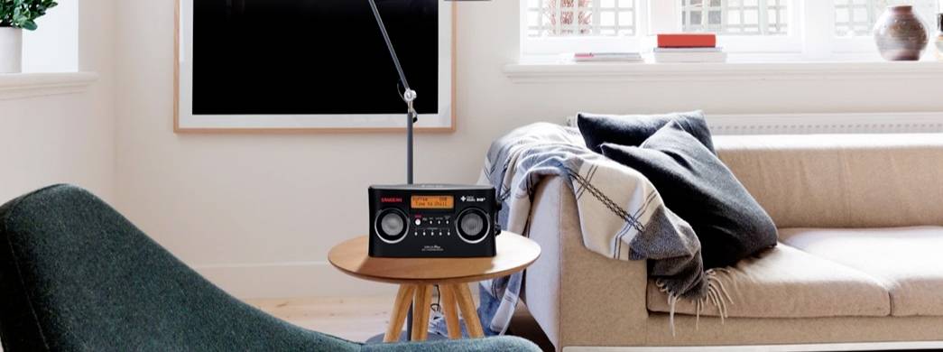 Un salon douillet avec des fauteuils, une table et une radio au centre. À l'arrière-plan, une grande fenêtre laissant entrer beaucoup de lumière du jour.