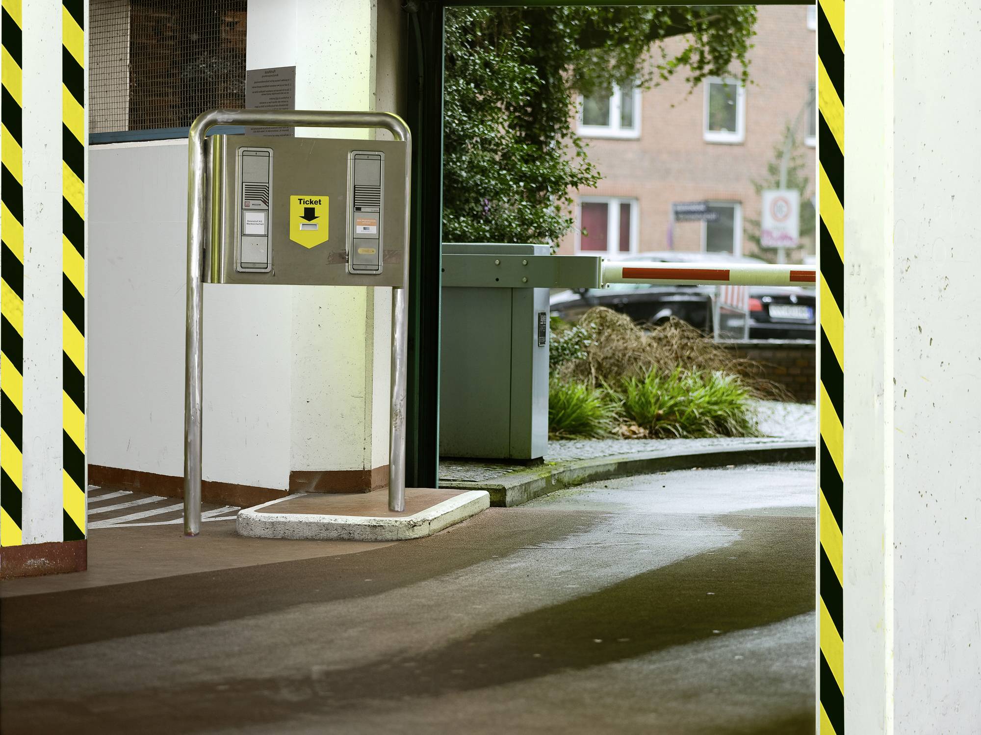 Entrée de parking avec barrière et distributeur de tickets. Des arbres et des bâtiments en briques en arrière-plan. Panneau de signalisation visible.