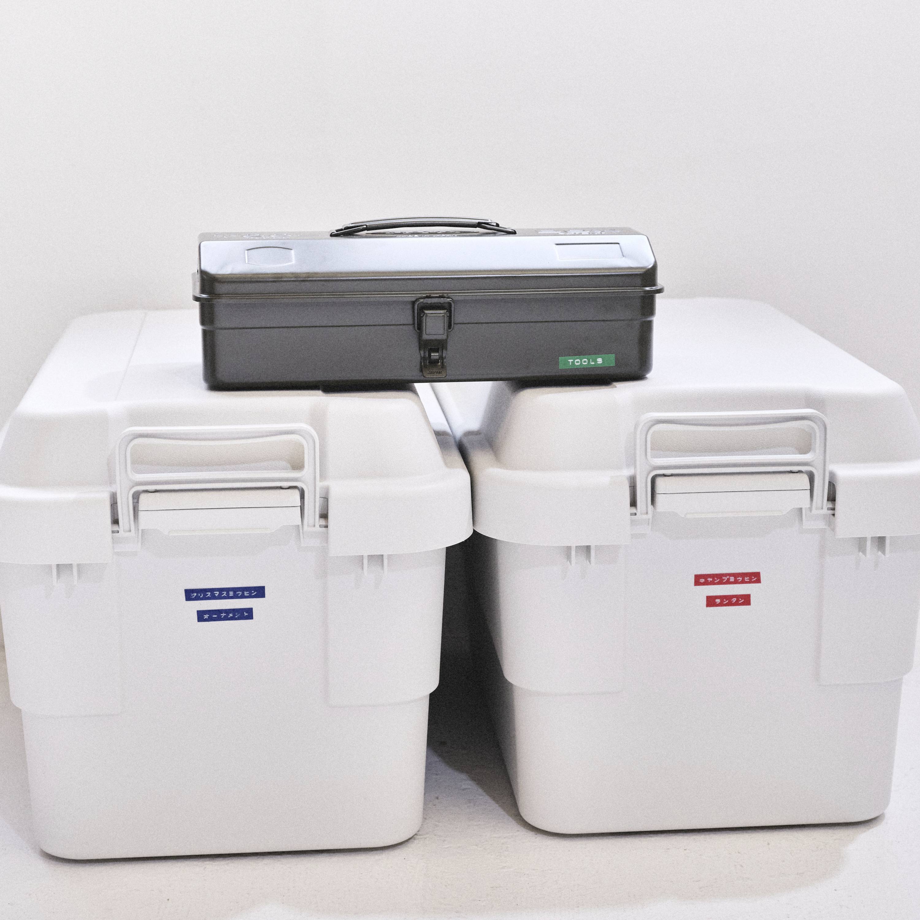 Une boîte à outils noire est posée sur deux caisses blanches avec des étiquettes en bleu et en rouge, placées devant un mur blanc.