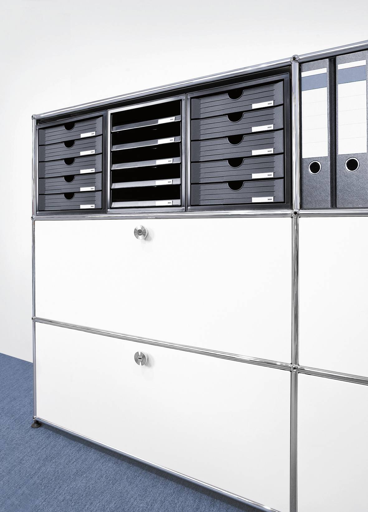 Un classeur blanc avec des compartiments et des dossiers sur un sol bleu. Il contient des tiroirs noirs et des chemises blanches avec des étiquettes.