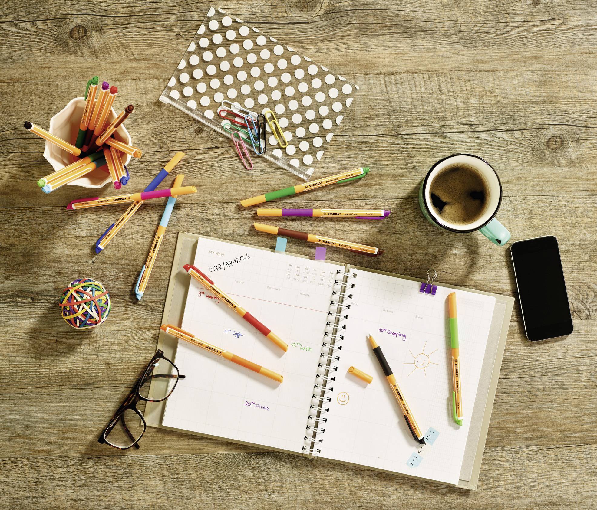 Un agenda ouvert avec des stylos colorés posé dessus est placé sur une table en bois à côté de lunettes, d'un téléphone portable, d'une tasse de café et d'un pot à crayons.